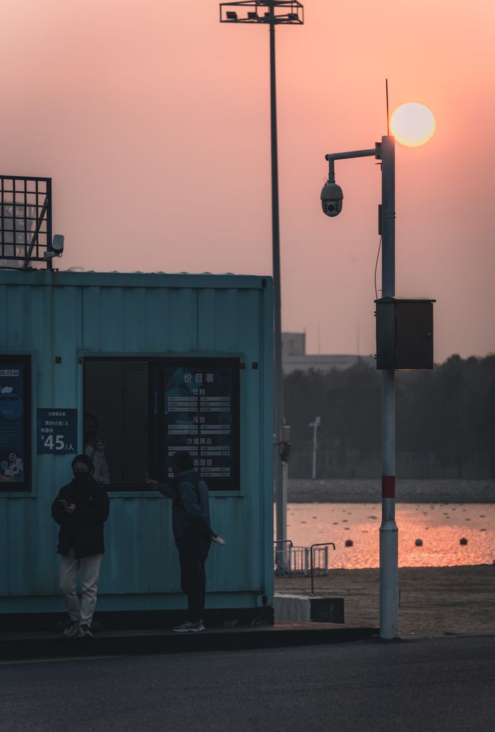 Silhouettes of people by a waterfront during sunset, with an urban ambiance and calm water.